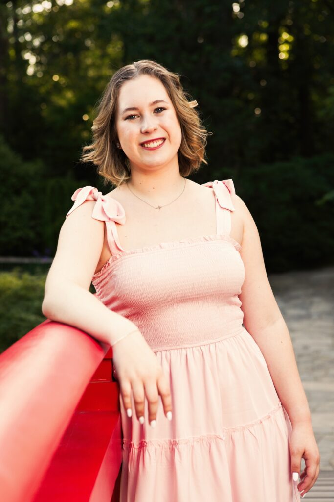 Senior girl wearing a light pink dress, posed by a red bridge at a local garden. Durham NC Senior Portrait Locations at a beautiful public garden.